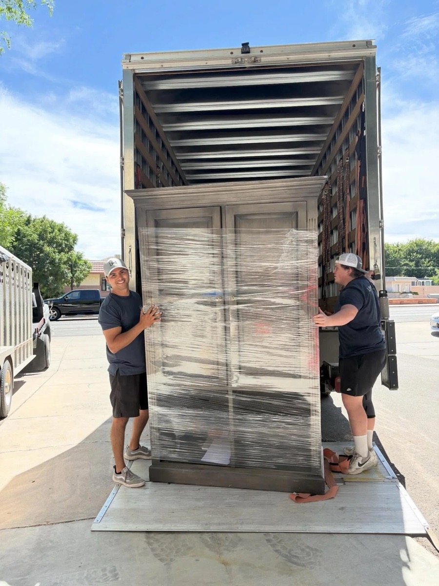Two movers loading a shrink-wrapped cabinet onto the truck lift